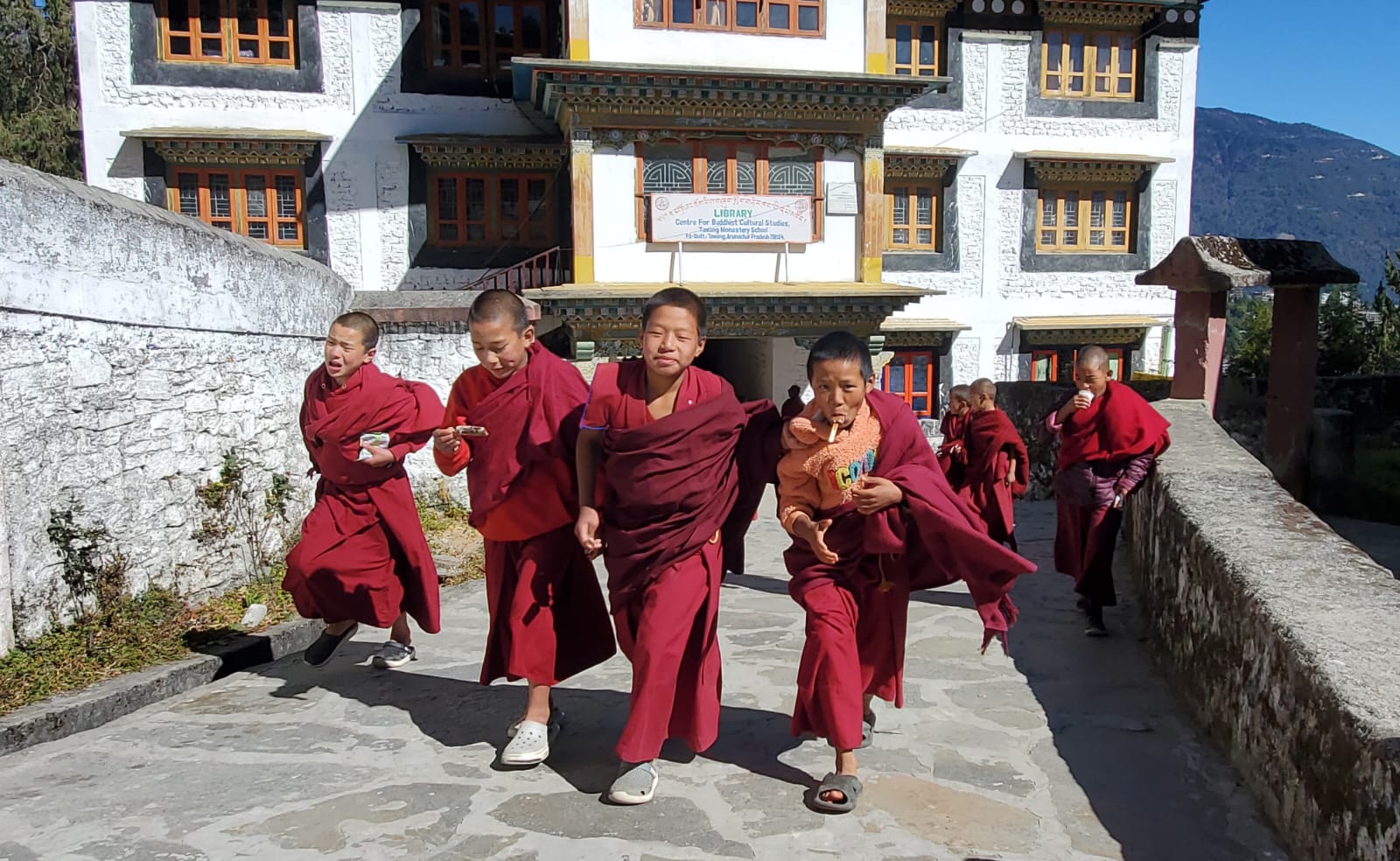 Young novice monks in maroon robes walking past the library at Tawang Monastery school