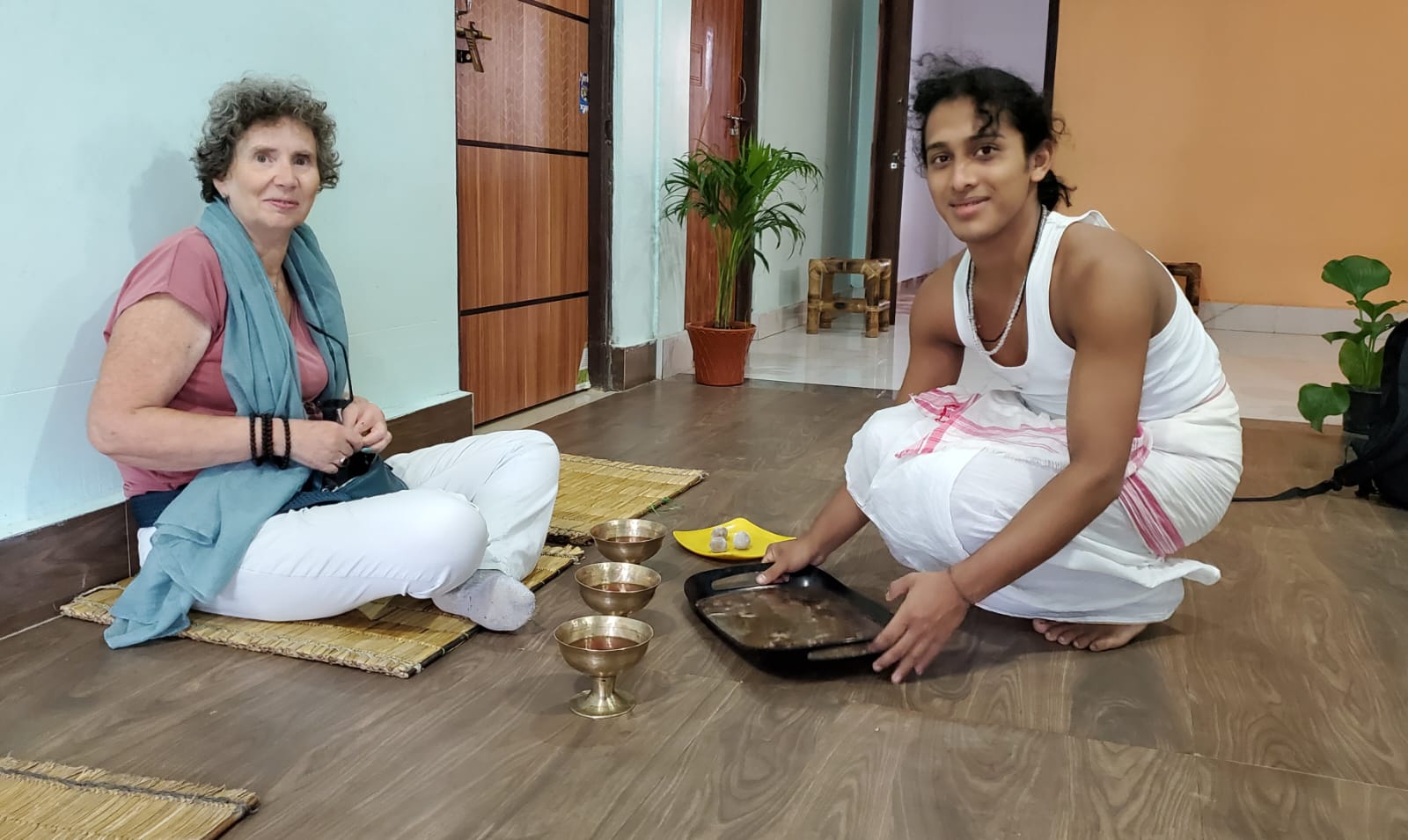 A young Vaishnavite monk in white dhoti preparing offerings beside a traveller at a Majuli satra