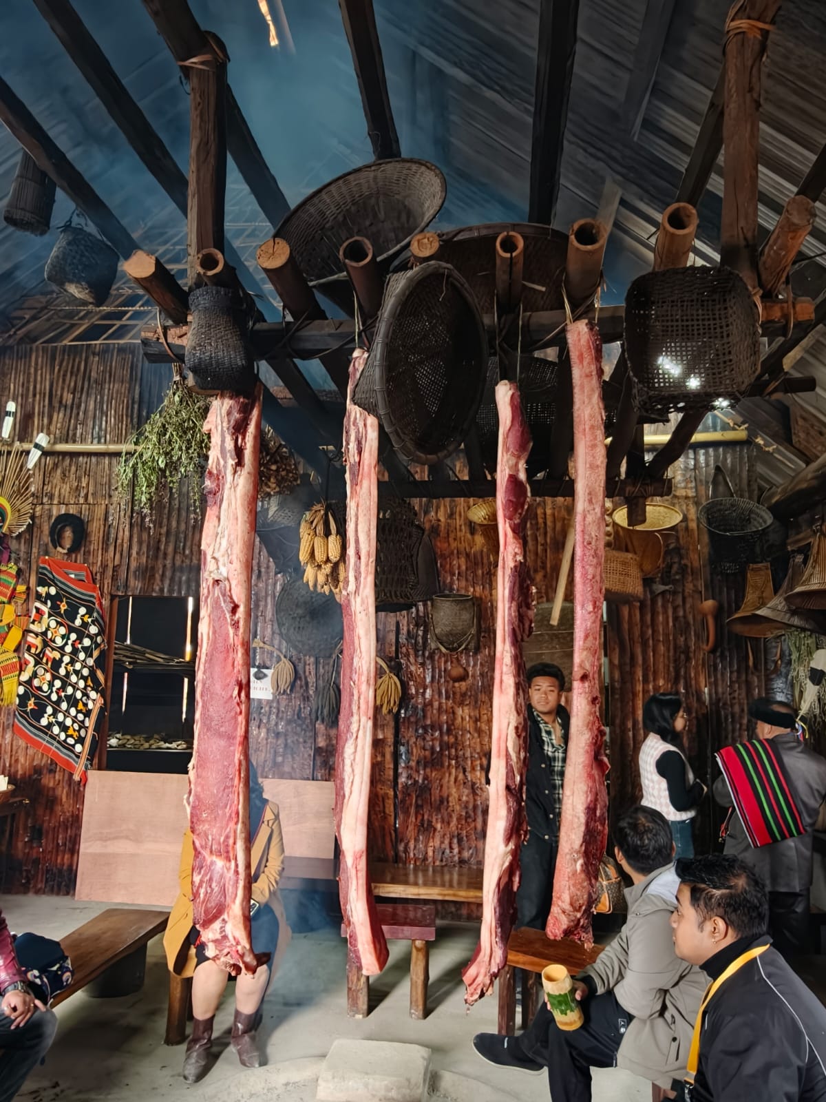 Cuts of meat hung from a beam beside cane baskets inside a timber and bamboo morung at Kisama