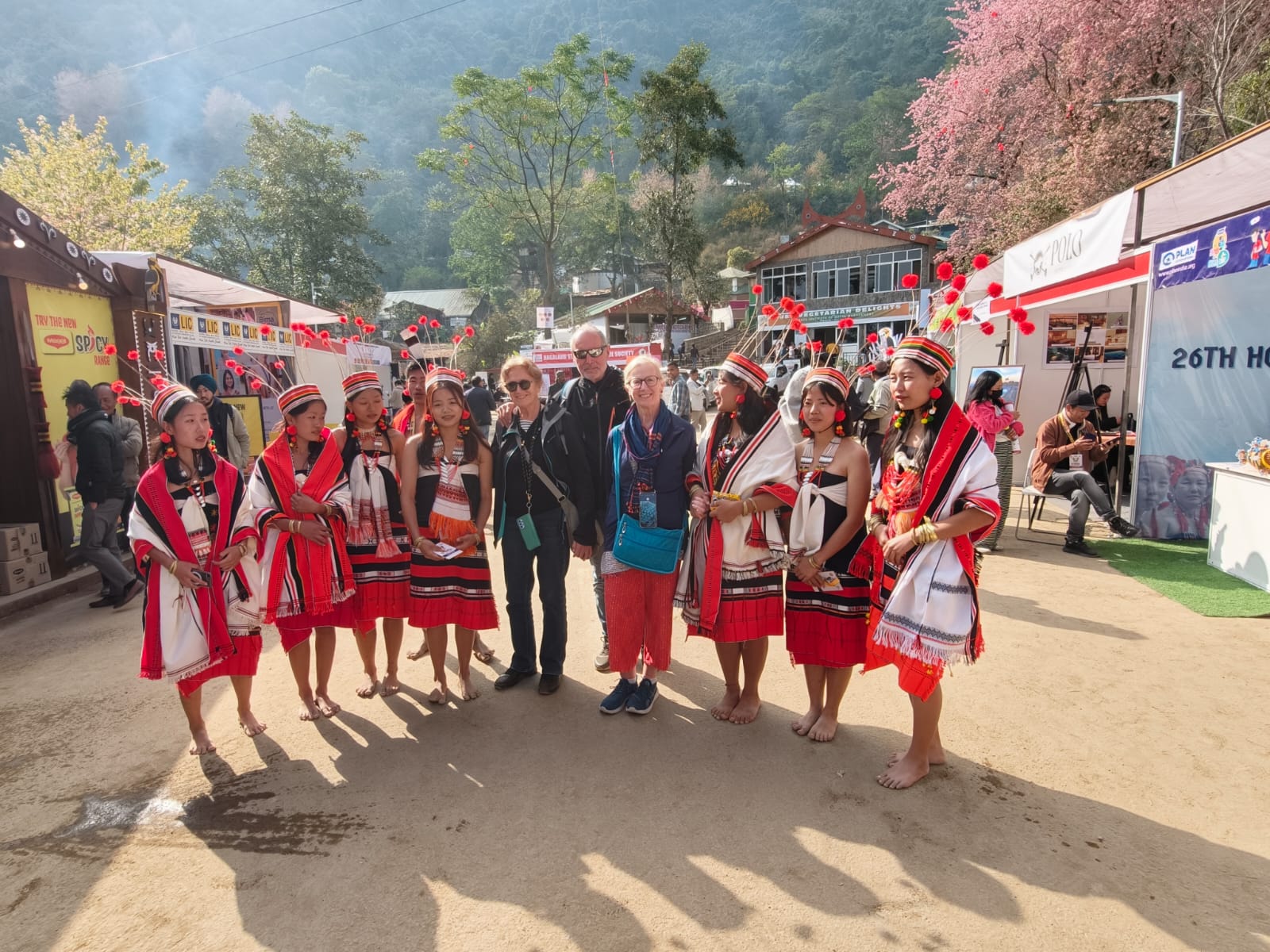Travellers standing with young Naga women in red and white traditional shawls at the Hornbill Festival, Kisama