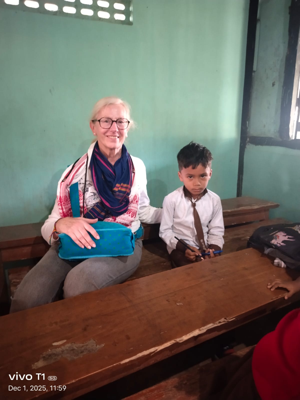 A traveller seated at a wooden desk beside a young boy in checked school uniform in an Assam village classroom
