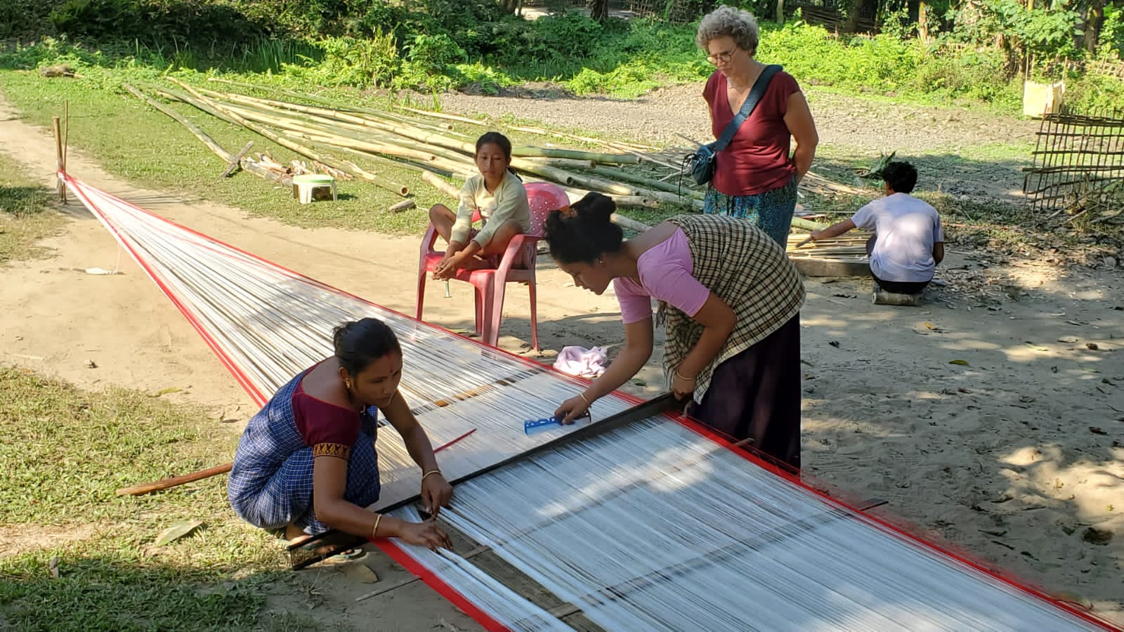 Assamese women preparing a warp on a ground loom while a traveller watches in a village in Assam