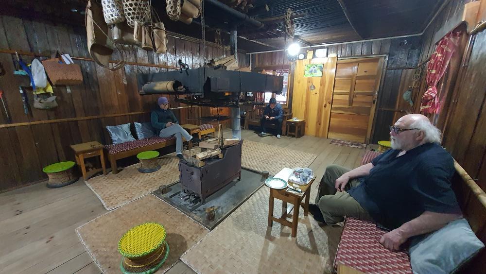 Jeans Pierre seated with hosts around the central hearth inside a traditional timber home near Aalo