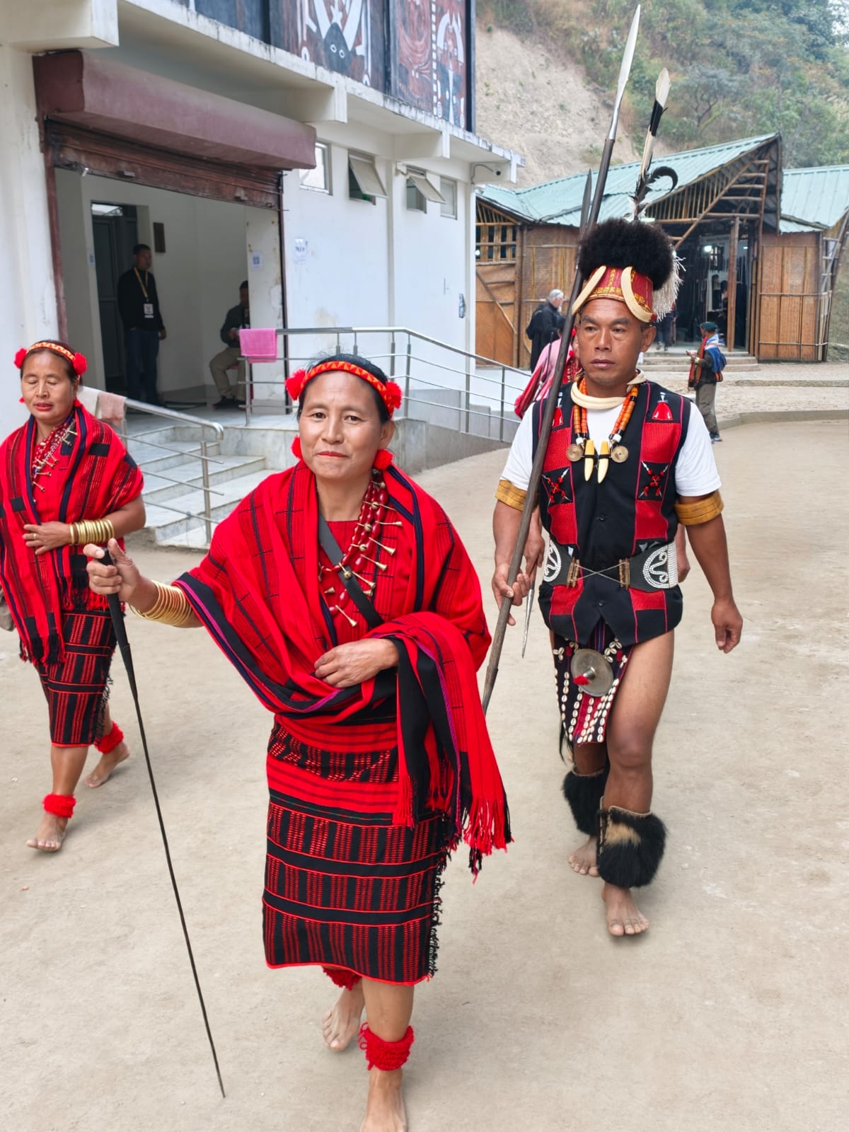 Two women in red and black woven wraps and a man carrying a spear walking through Kisama Heritage Village