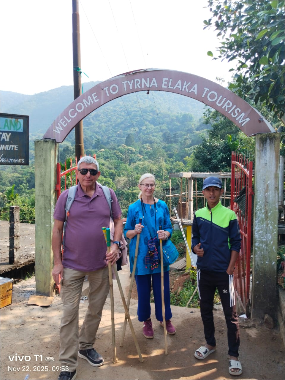 Two travellers with walking sticks under the Welcome to Tyrna Elaka arch at the start of the root bridge trail in Meghalaya