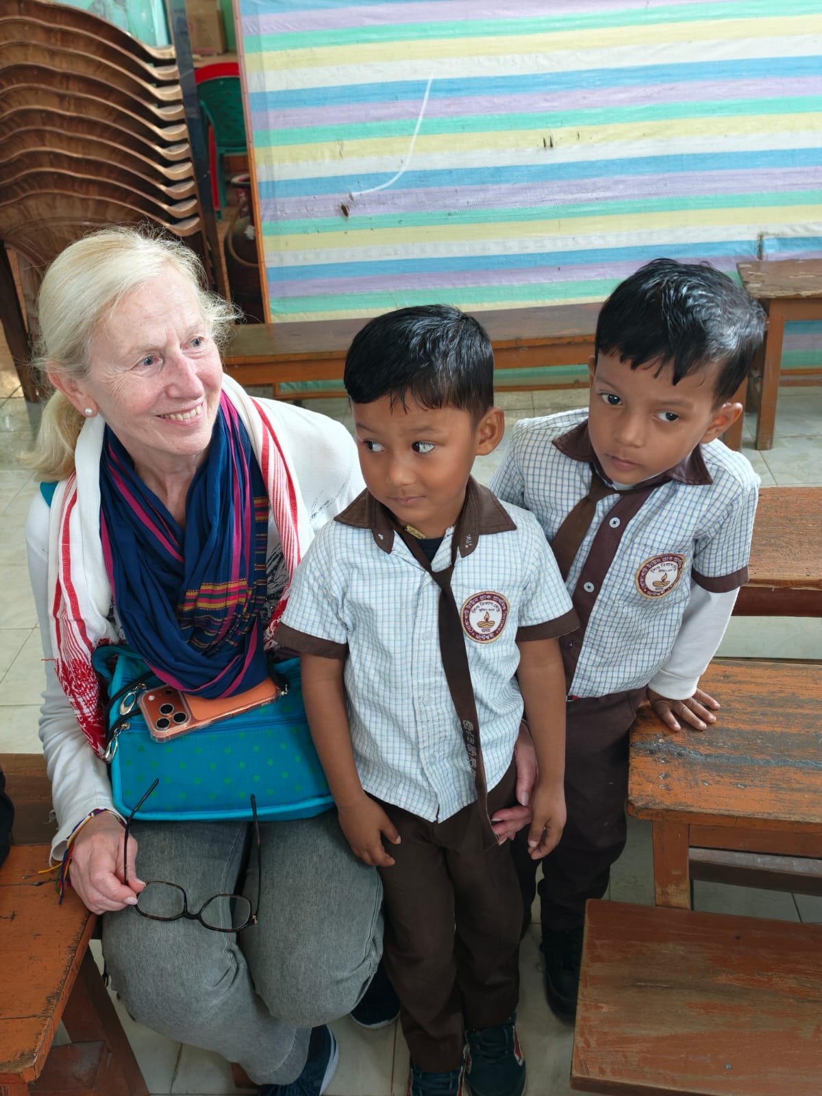A traveller wearing a gamosa seated with two schoolboys in uniform in a village classroom in Assam