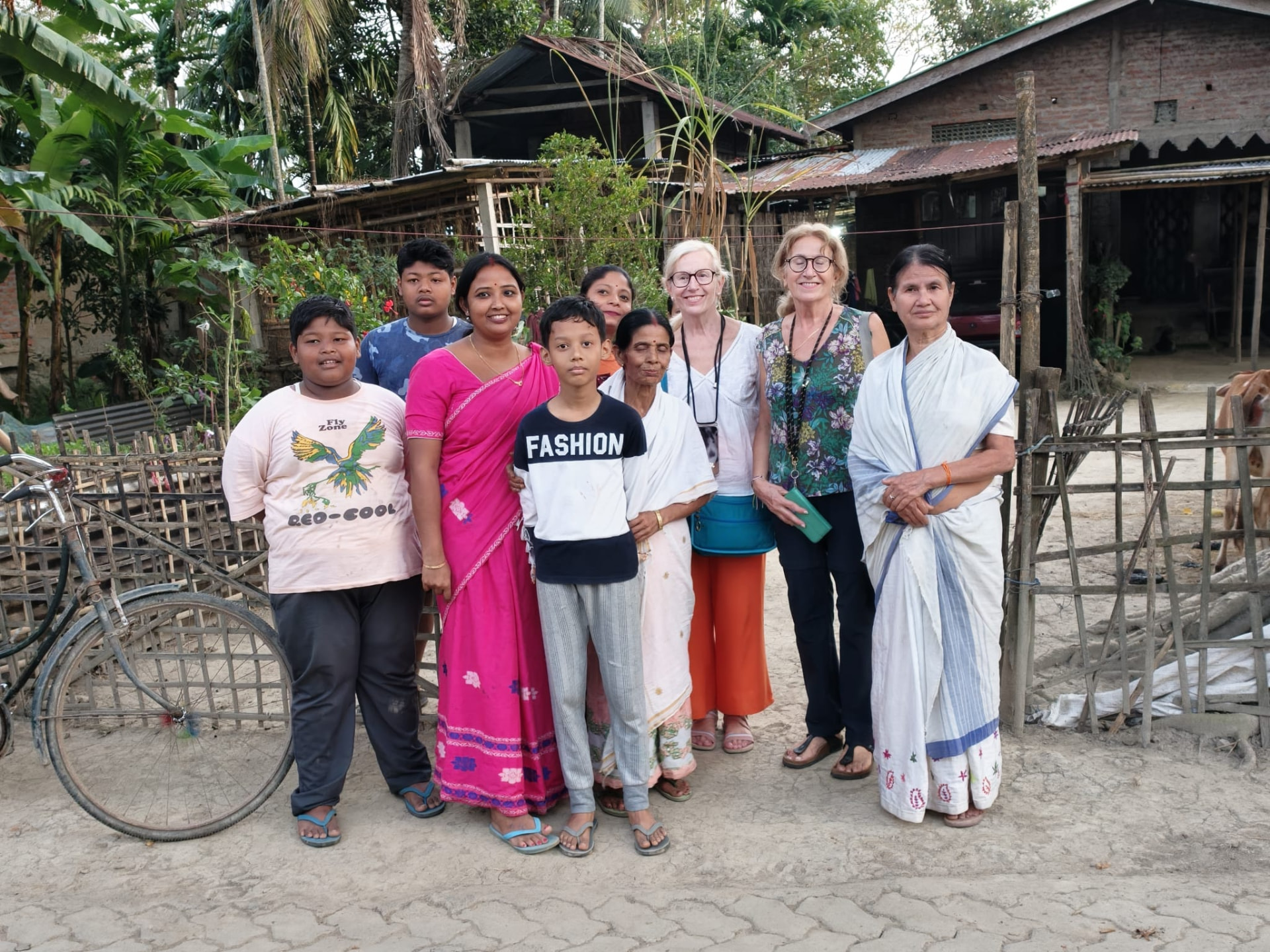 Travellers standing with an Assamese village family beside a bamboo fence in front of their home
