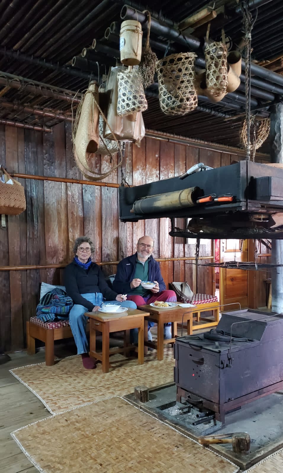 Robert and Odile eating in a traditional Monpa timber house with bamboo baskets hung above the hearth