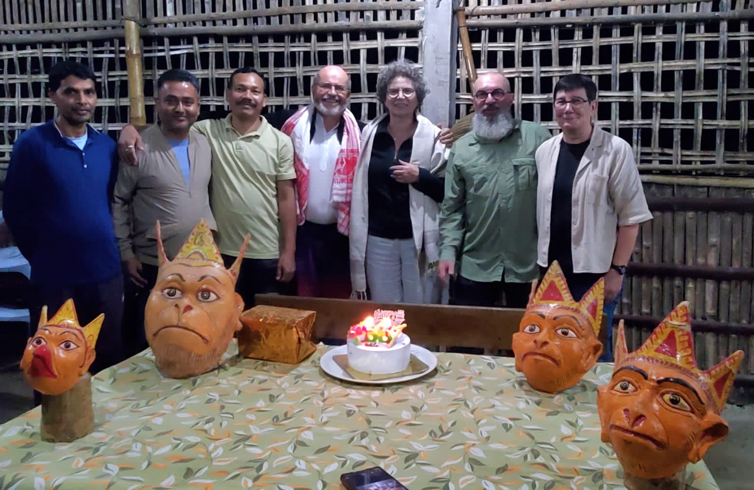 Group photograph with guides behind a birthday cake and painted Assamese masks in a bamboo hall on Majuli