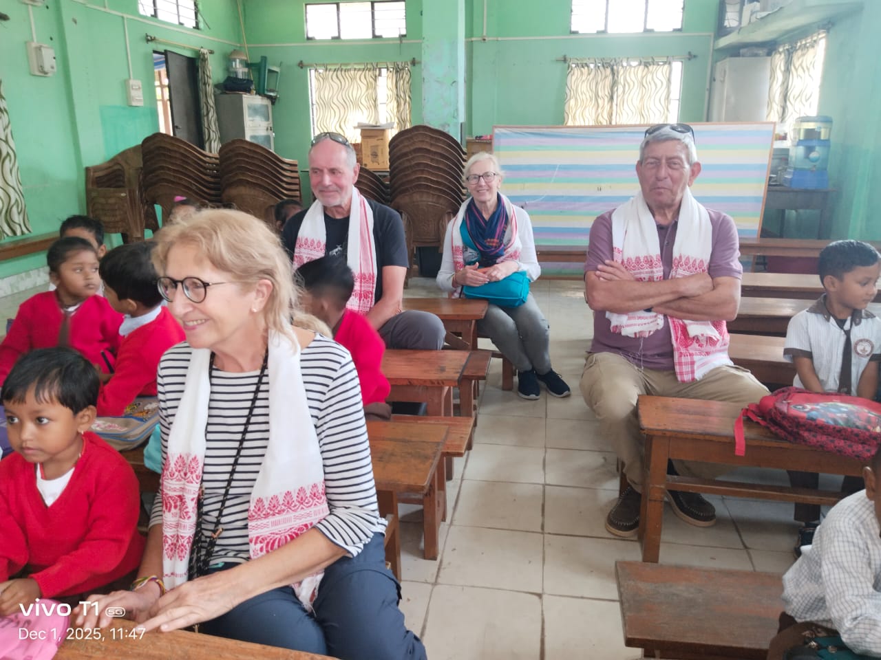 Four travellers wearing red and white gamosa scarves seated among schoolchildren in a green village classroom in Assam